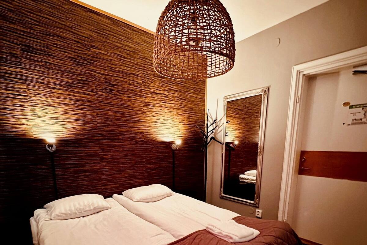 Un lit bien fait, avec des draps blancs et une serviette pliée, un luminaire en fibres naturelles au-dessus et un miroir au mur.