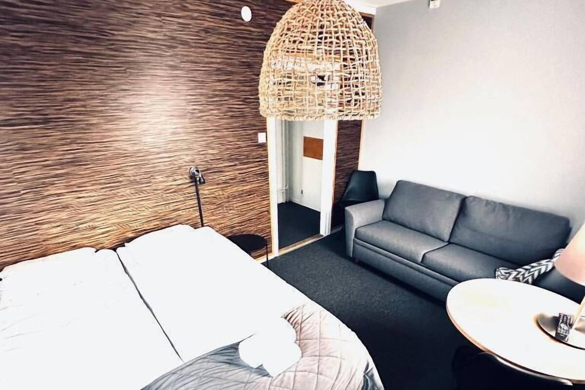 Une chambre d’hôtel moderne avec un lit, un canapé gris, une table ronde et un luminaire tressé.