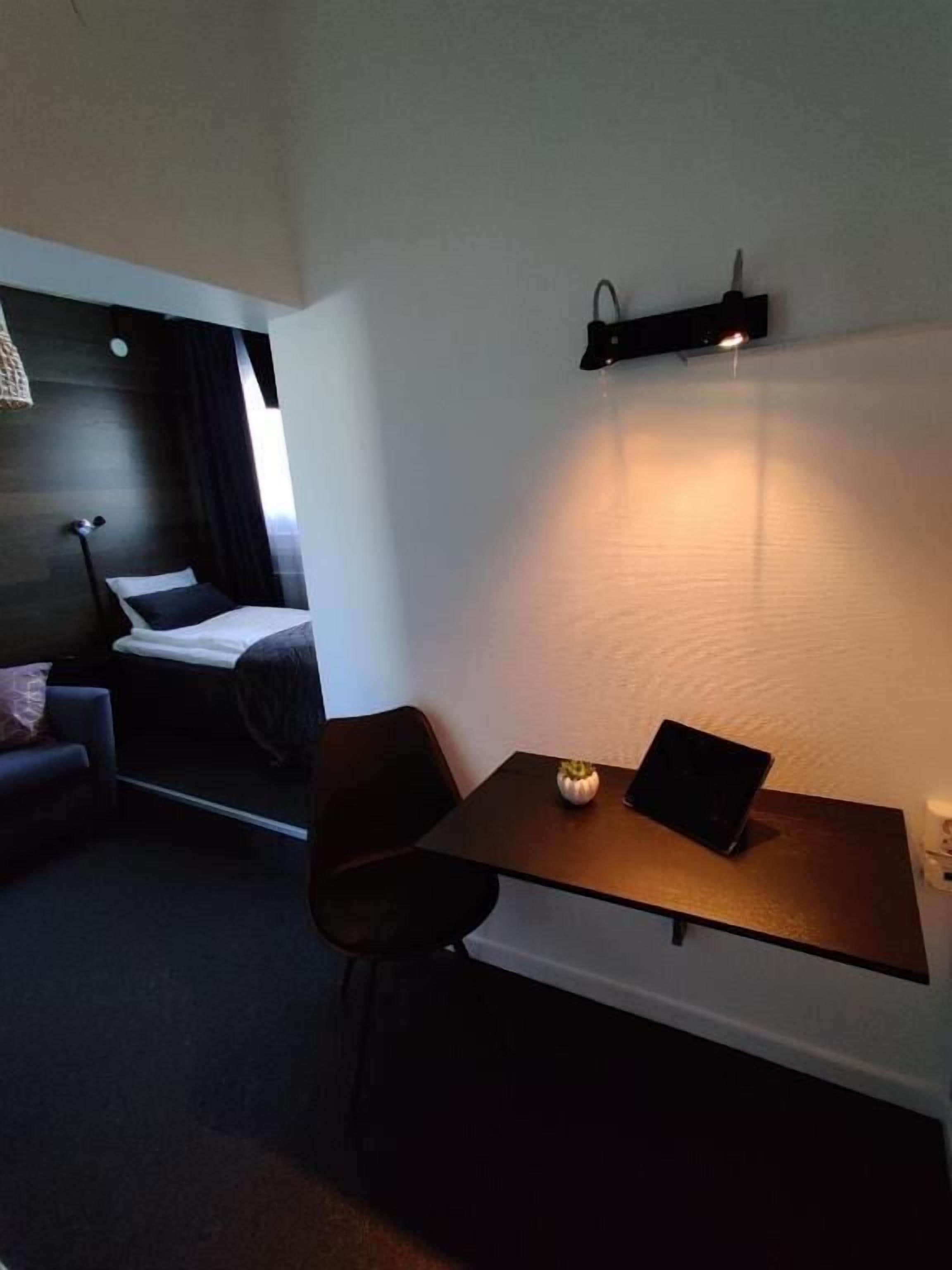 Habitación de hotel pequeña con una cama, una mesita con una computadora portátil, una silla y una lámpara de noche.