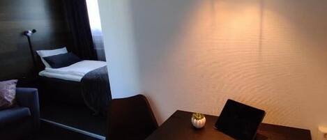 Habitación de hotel pequeña con una cama, una mesita con una computadora portátil, una silla y una lámpara de noche.