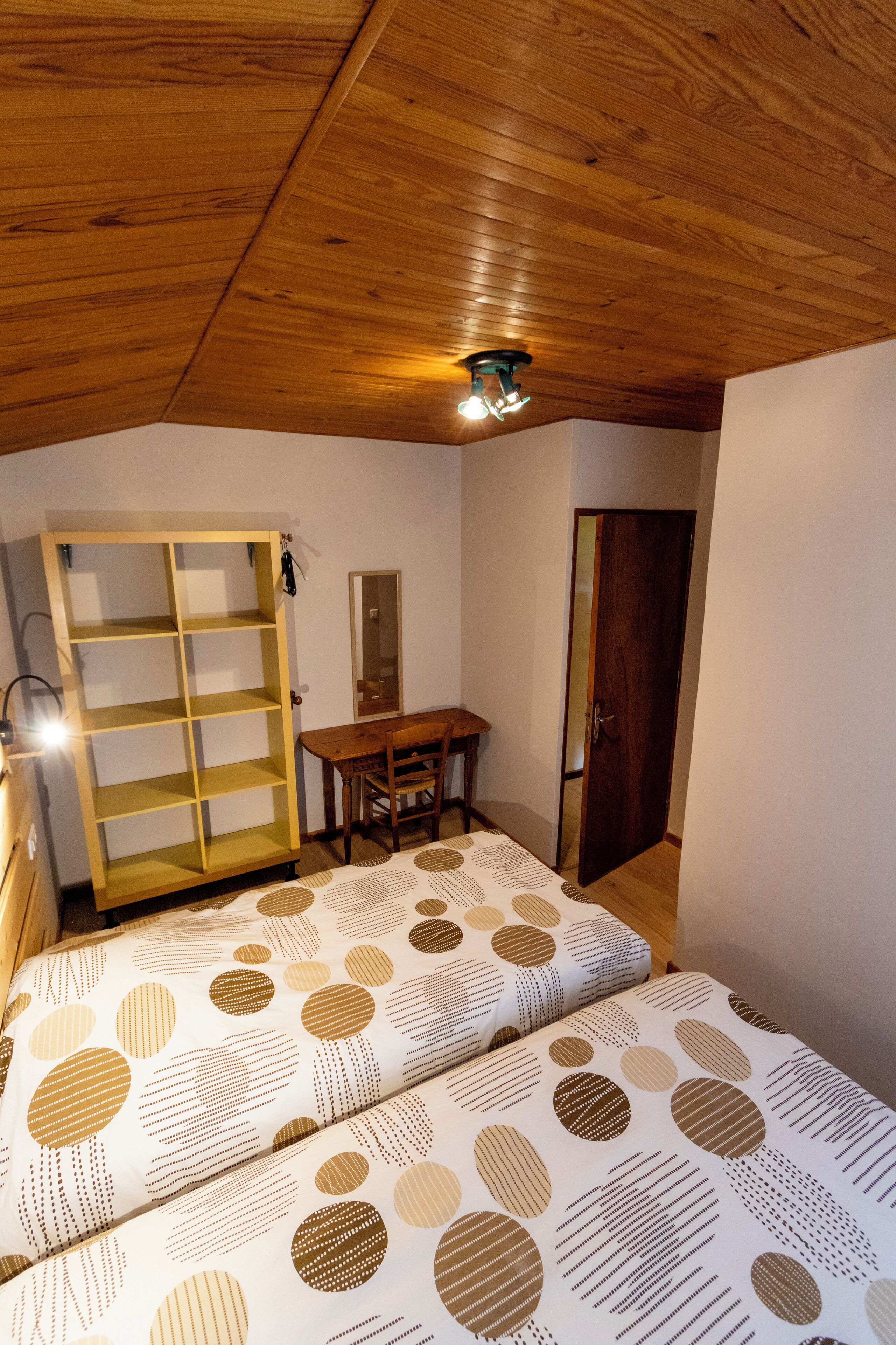 A bedroom with a wooden ceiling, a bed with patterned bedding, a wooden desk, a mirror, and a wardrobe.