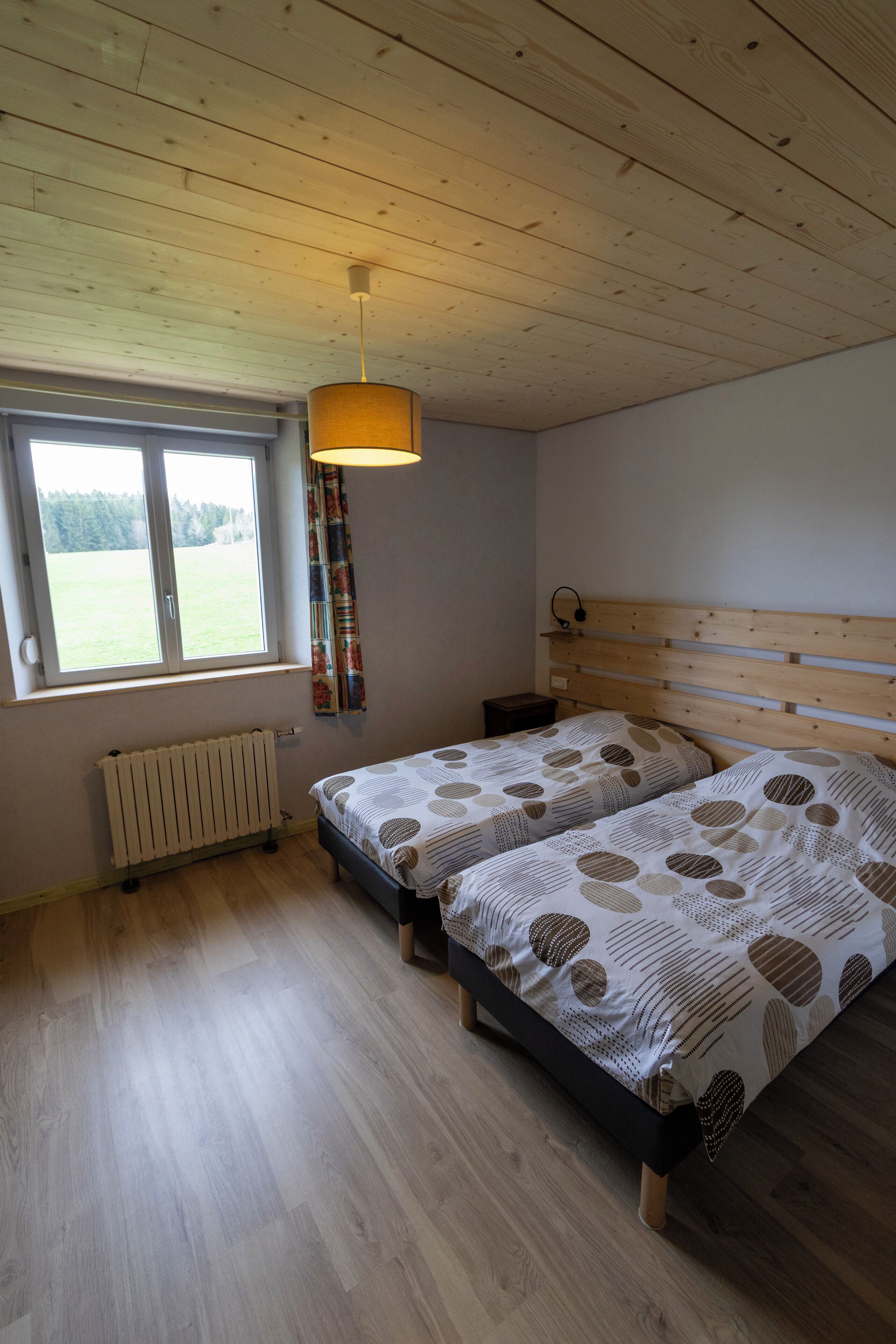 A bedroom with two beds, a wooden ceiling, a window with a view of a field, and a radiator.