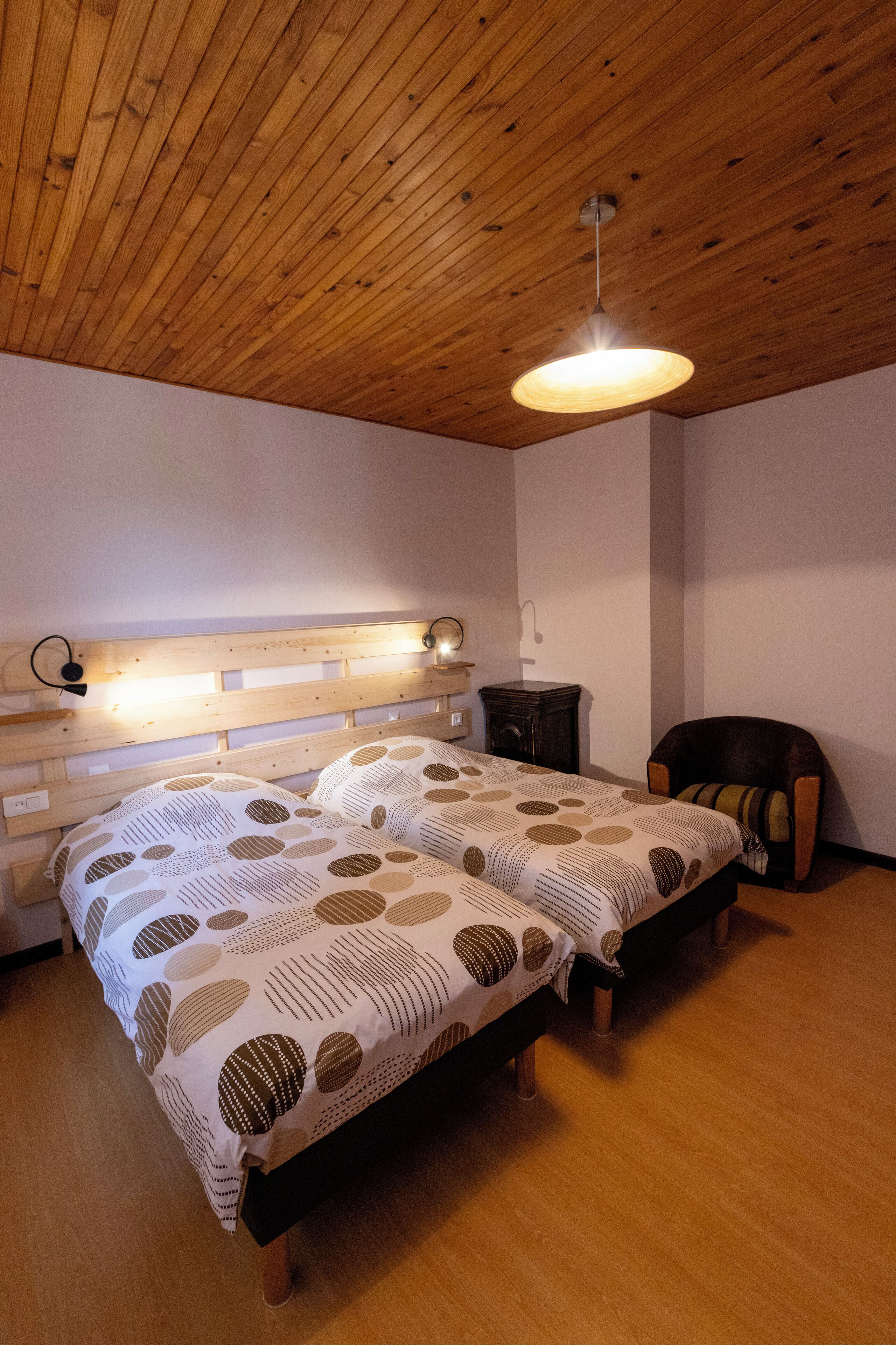 A bedroom with a wooden ceiling, two beds with patterned bedding, a nightstand, and a chair.