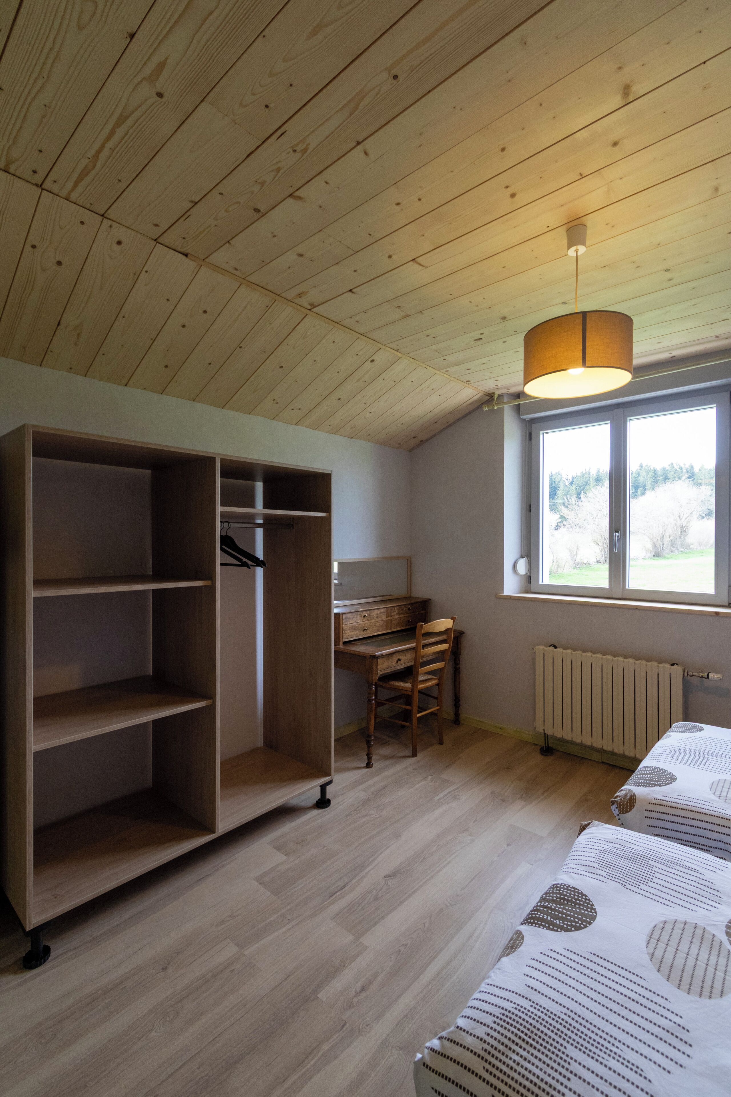 Een kamer met een houten plafond, een grote inbouwkast, een bureau met stoel, een radiator en een raam met uitzicht op bomen.