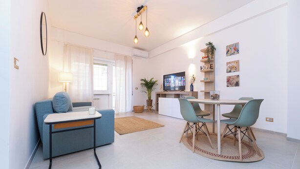 A modern living room with a sofa, a dining table with chairs, a TV, and a kitchen area with a fridge and shelves.