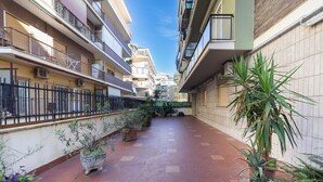 A narrow pedestrian pathway flanked by multi-story residential buildings with balconies, potted plants, and a tiled floor.