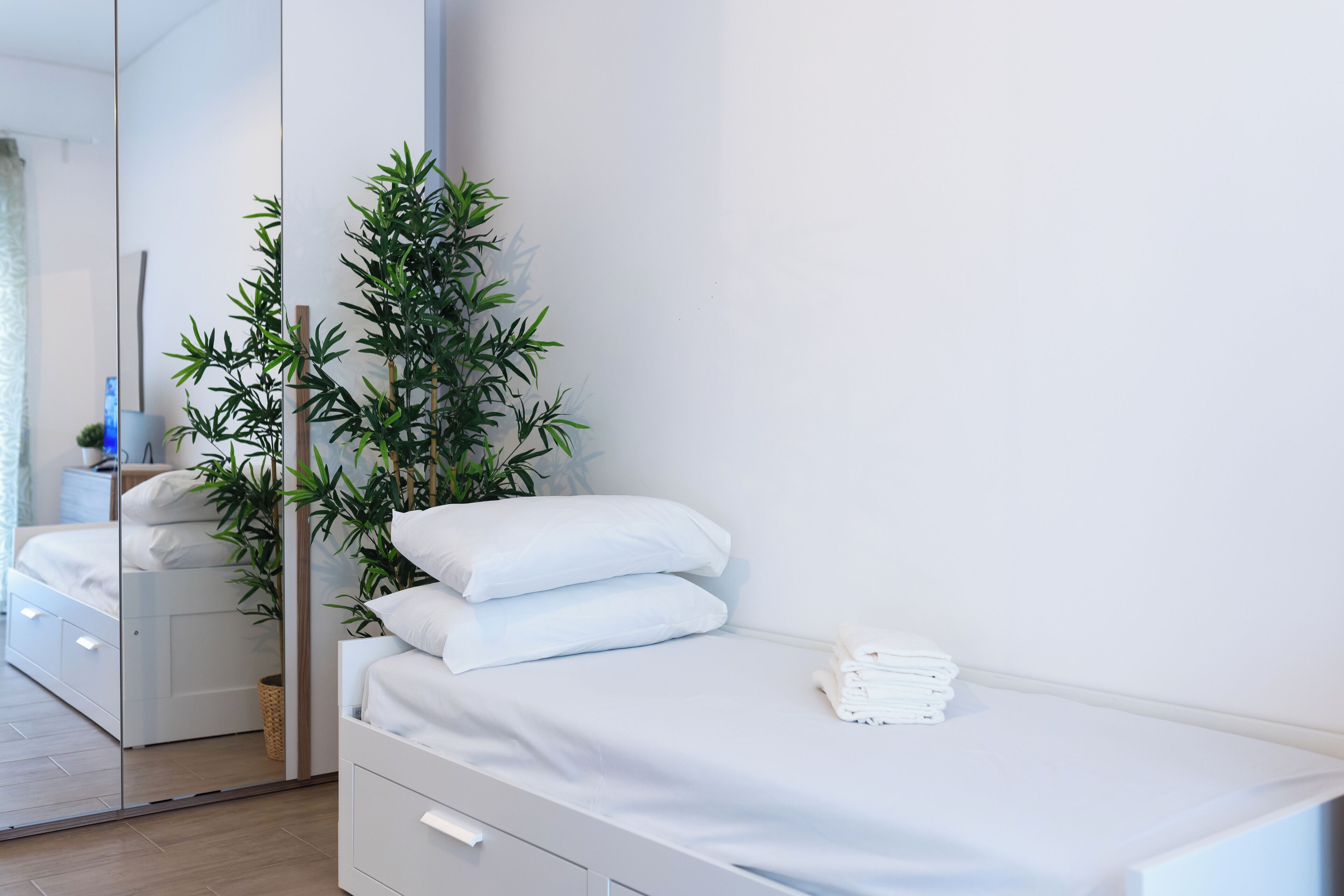 Un lit bien fait, avec du linge de lit blanc, une petite table de chevet surmontée d’une lampe blanche, et une grande plante verte placée à côté du lit.