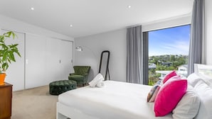 Une chambre moderne avec un grand lit, un fauteuil vert, un lampadaire et une vue sur la ville par une grande fenêtre.