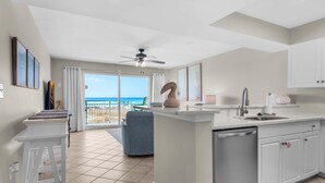 A modern kitchen with a central island, a dining area with a blue chair, and a view of the ocean through large windows. - Pelican Isle 117 (Fort Walton Beach)