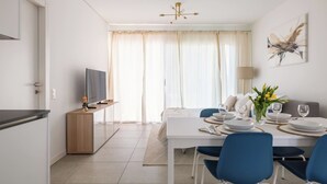 A modern dining area with a white table set for four, blue chairs, and a television on a wooden stand. - Cozy & Modern Flat for 6 people / Free Parking (Lugano)