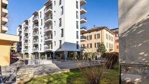 A multi-story residential building with balconies, a paved courtyard, and surrounding greenery. - Cozy & Modern Flat for 6 people / Free Parking (Lugano)