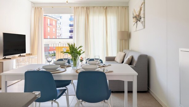 A modern living room with a dining area, a flat-screen TV, and a view of the cityscape. - Cozy & Modern Flat for 6 people / Free Parking (Lugano)