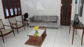 Un salon avec un canapé gris, des chaises en bois, une table basse ornée d’un vase et un ventilateur.