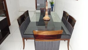 Une table à manger avec un plateau en verre et des chaises en bois, entourée d’un sol carrelé blanc et agrémentée d’une plante en pot.
