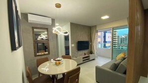 A modern living room with a grey sofa, a dining table with chairs, a flat-screen TV, and a large window offering a city view. - Edificio Promenade II - Apt 604 (Maceio)