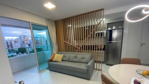 A modern living room with a grey sofa, a wooden partition, a dining table with chairs, and a kitchen area with a refrigerator. - Edificio Promenade II - Apt 604 (Maceio)
