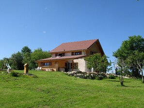Une maison à deux étages, avec un toit en pente, des fondations en pierre et une cour couverte d’herbe.