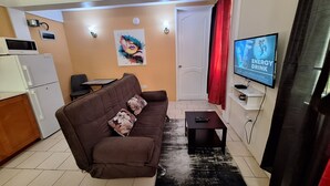 A compact living space with a brown sofa, a small black table, a TV on the wall, and a kitchen area with a refrigerator and microwave.