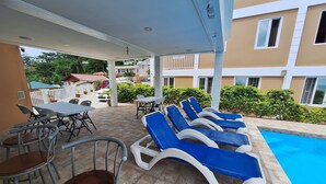 A covered outdoor area with lounge chairs and tables, a swimming pool, and a building in the background.