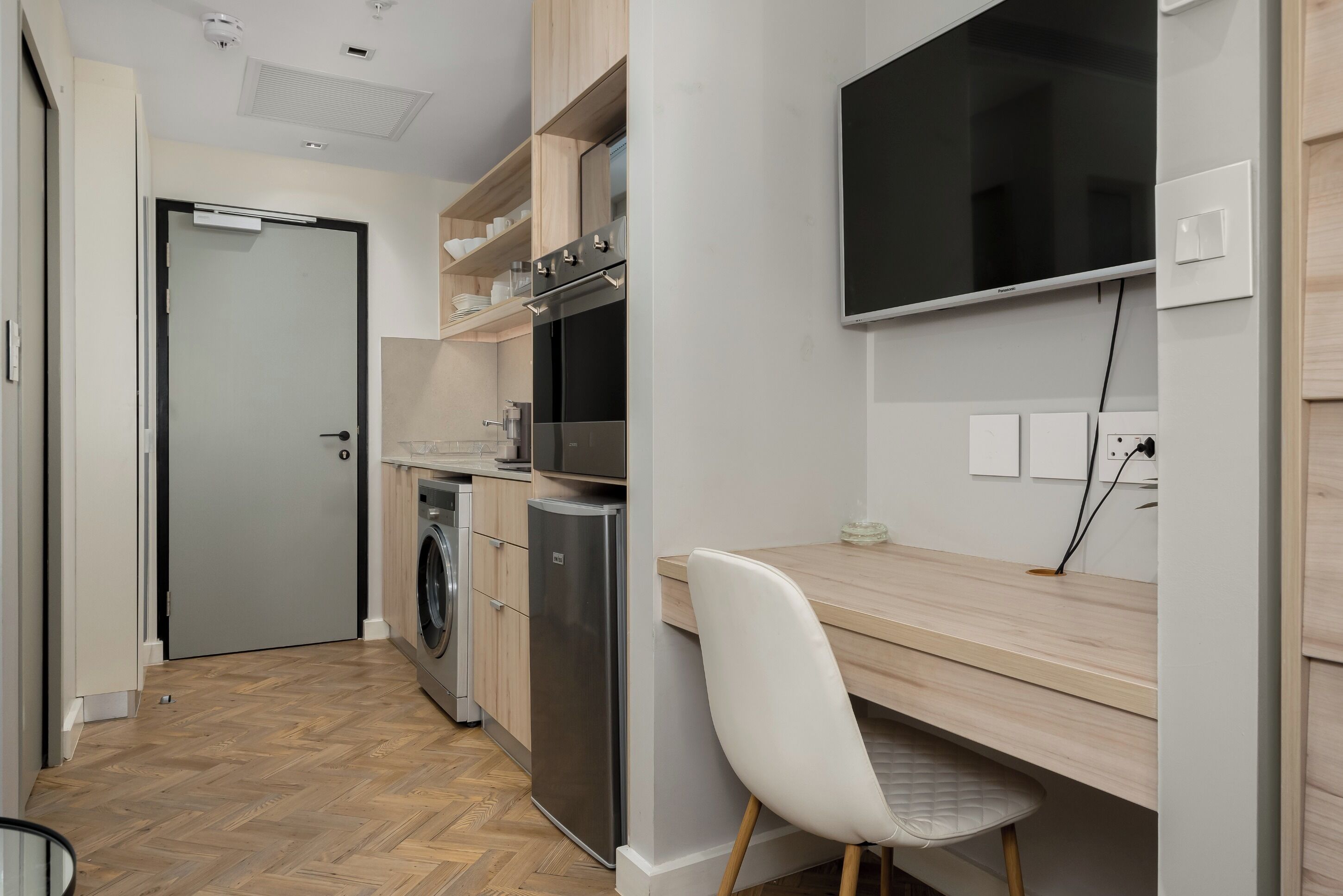 A modern kitchen with a built-in oven, microwave, and dishwasher. There is a wooden dining table with a white chair and a flat-screen TV mounted on the wall.