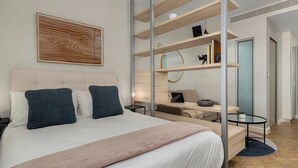 Une chambre à coucher moderne avec un grand lit, une tête de lit, une table de chevet, un bureau, une chaise, une bibliothèque et une œuvre d’art accrochée au mur.