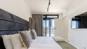 A modern hotel room with a large bed, a view of the city through the window, and a flat-screen TV on the wall. - The Sage | Modern and Bright Sea Point Apartment (Cape Town)