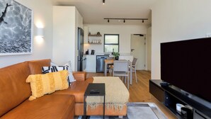 A modern living room with a brown leather sofa, a flat-screen TV, and a dining area with a kitchen in the background. - The Sage | Modern and Bright Sea Point Apartment (Cape Town)