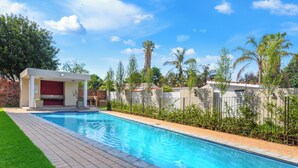 A swimming pool with a brick-lined deck, a covered seating area, and a fence with greenery.