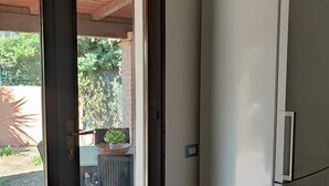 A room with a white refrigerator, a door leading to an outdoor area with a table and chairs, and a potted plant. - Charming 2-bedroom cottage with WiFi, AC in lovely Quartu Sant'Elena (Quartu Sant'Elena)
