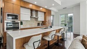 A modern kitchen with wooden cabinets, a large island with bar stools, a built-in oven, and a view of the city through large windows. - Central CN Tower View | 3bed 3 bath | 6 Guest | Pool | Gym | Parking (Toronto)