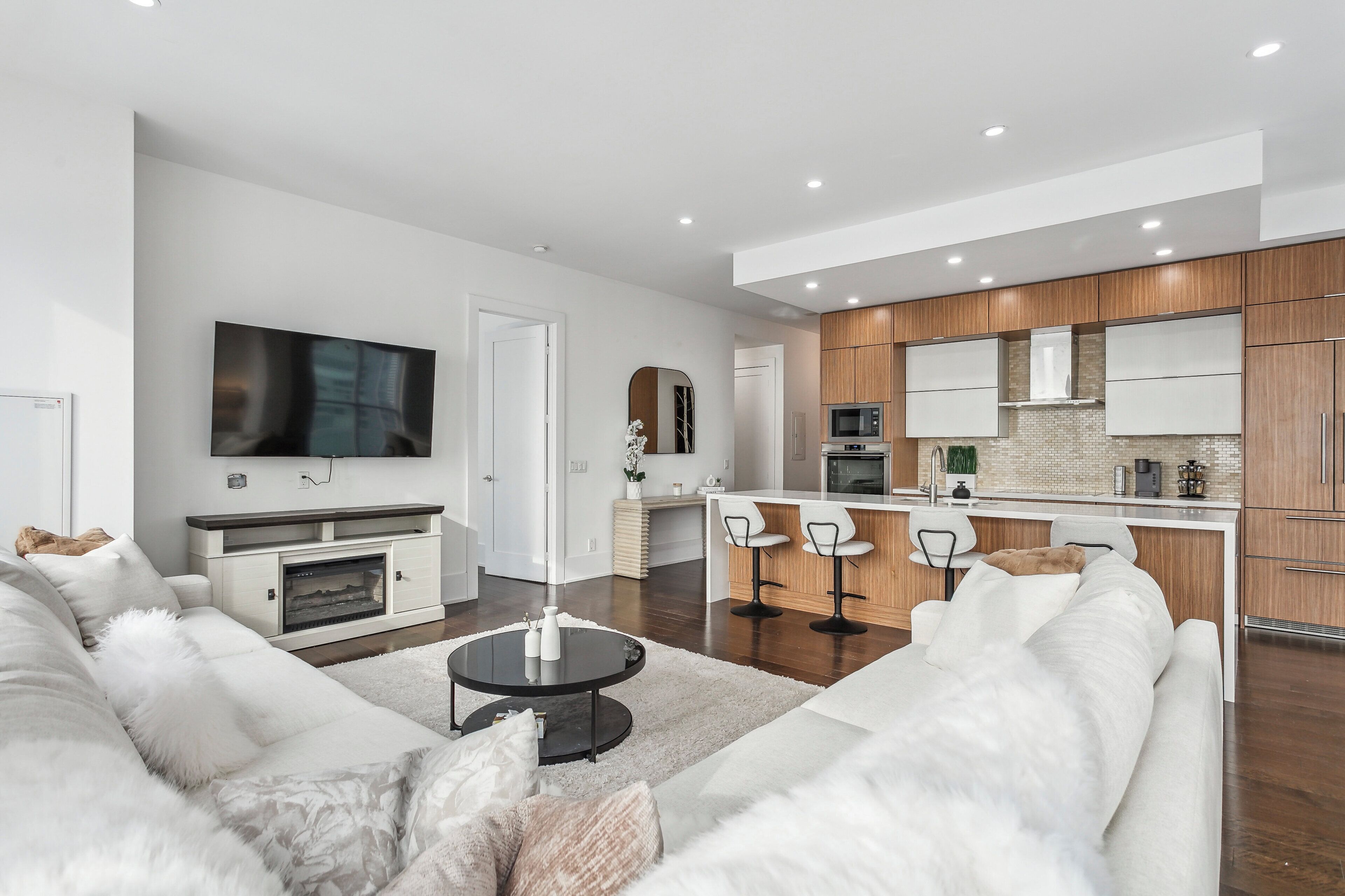A modern living room with a sofa, a coffee table, and a TV. There is a kitchen area with wooden cabinets and a bar.