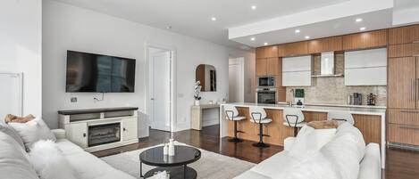 A modern living room with a sofa, a coffee table, and a TV. There is a kitchen area with wooden cabinets and a bar.