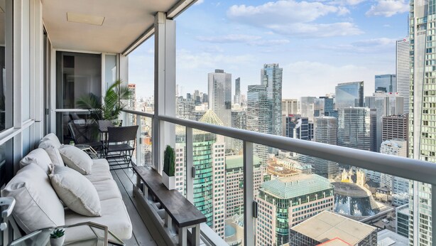 A balcony with a sofa, chairs, and a small table, overlooking a cityscape with modern buildings. - Central CN Tower View | 3bed 3 bath | 6 Guest | Pool | Gym | Parking (Toronto)