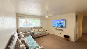 Ein Wohnzimmer mit einer Couch, einem an der Wand befestigten Fernseher und einem Fenster mit Jalousien.