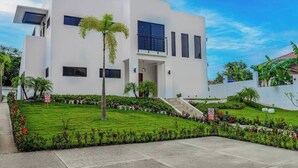 Une maison moderne à deux étages, avec une façade blanche, un petit jardin devant agrémenté d’un palmier et un escalier menant à l’entrée.