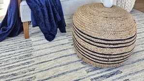A living room with a grey sofa, patterned cushions, a blue throw blanket, a woven ottoman, and a striped rug.