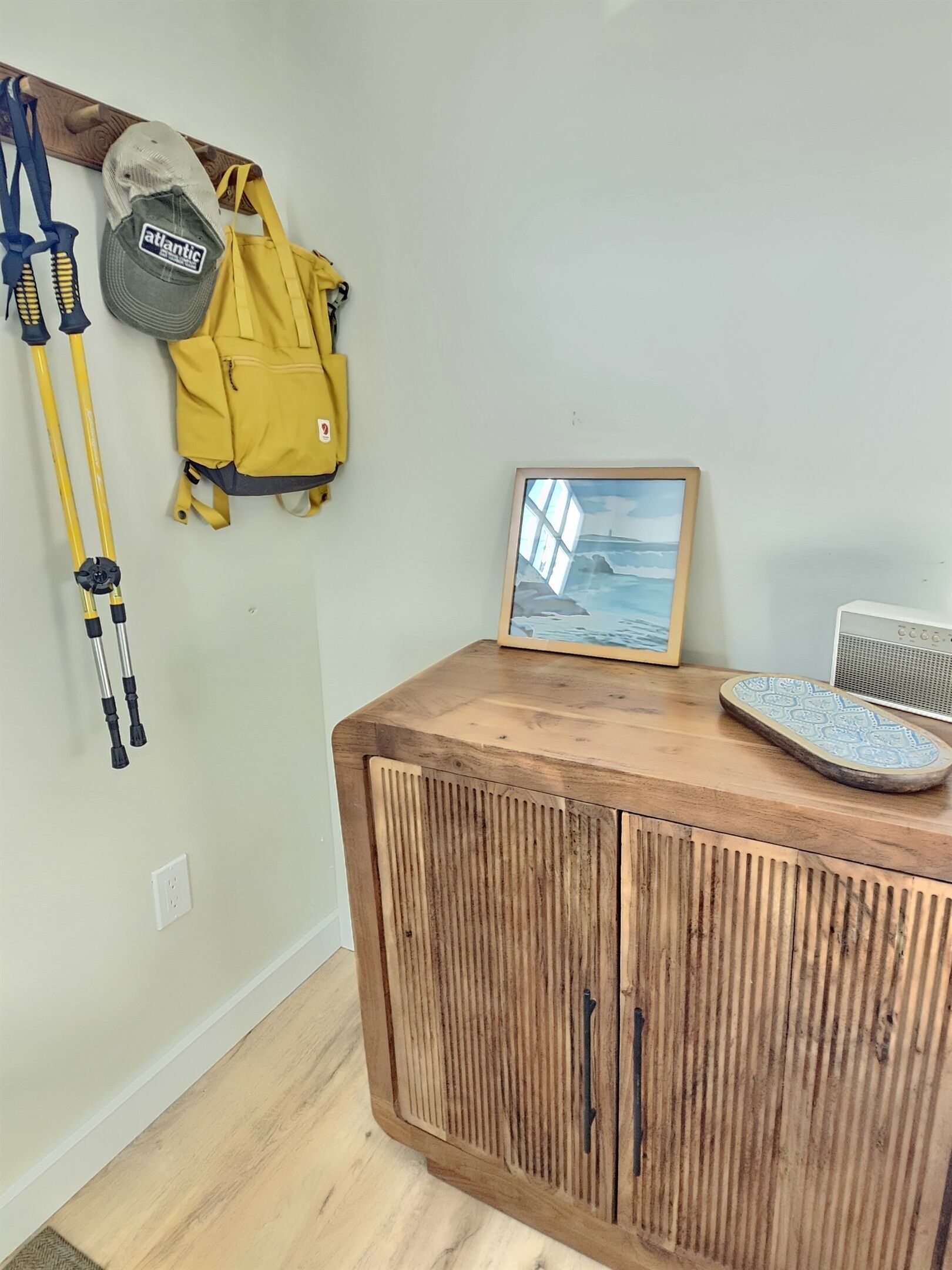 A wooden cabinet with a framed picture of a seascape, a yellow backpack, and hiking poles.