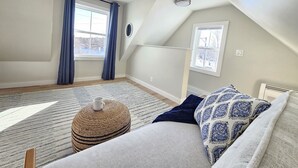 A cozy attic bedroom with a sloped ceiling, a large window with blue curtains, a round woven ottoman, a patterned throw pillow, and a ceiling fan.
