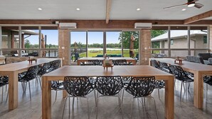 Een ruime eetruimte met een grote houten tafel en zwarte stoelen, grote ramen met uitzicht op de omgeving, en een plafondventilator.