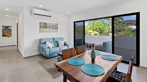 A modern living room with a blue sofa, a wooden dining table with green placemats, and a large sliding glass door leading to a balcony with a view of trees and buildings. - Casa Corales Dreams Deluxe (Playa Flamingo)