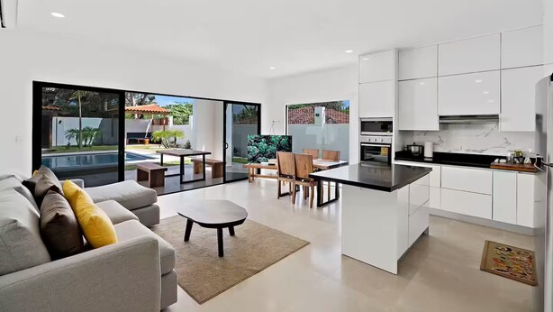 A modern kitchen with white cabinets, a dining area with a table and chairs, and a large sliding glass door leading to a pool. - Casa Corales Dreams Deluxe (Playa Flamingo)