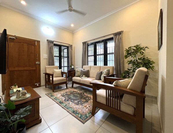 Un salon comprenant un canapé, deux fauteuils, une table basse et un téléviseur. Il y a des fenêtres avec des rideaux, un ventilateur de plafond et une plante en pot.