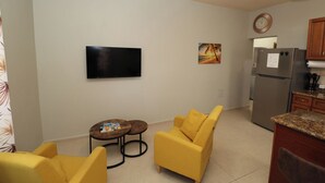 Een knus woongedeelte met een bank, een klein rond tafeltje, een flatscreen televisie aan de muur, een koelkast en een klok.