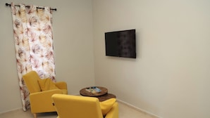 Een kamer met een gele fauteuil, een rond houten bijzettafeltje, een kunstwerk met een zwarte lijst aan de muur en een kamerbreed gordijn.