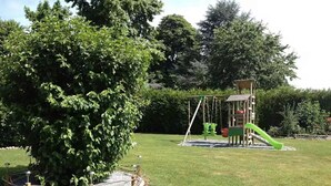 Un parque infantil con columpios y un tobogán, rodeado de césped cuidado y un árbol grande.