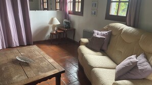 Una sala de estar acogedora con una mesa de madera, un sofá color crema con cojines, una pequeña mesita de noche con una lámpara y una ventana con cortinas.