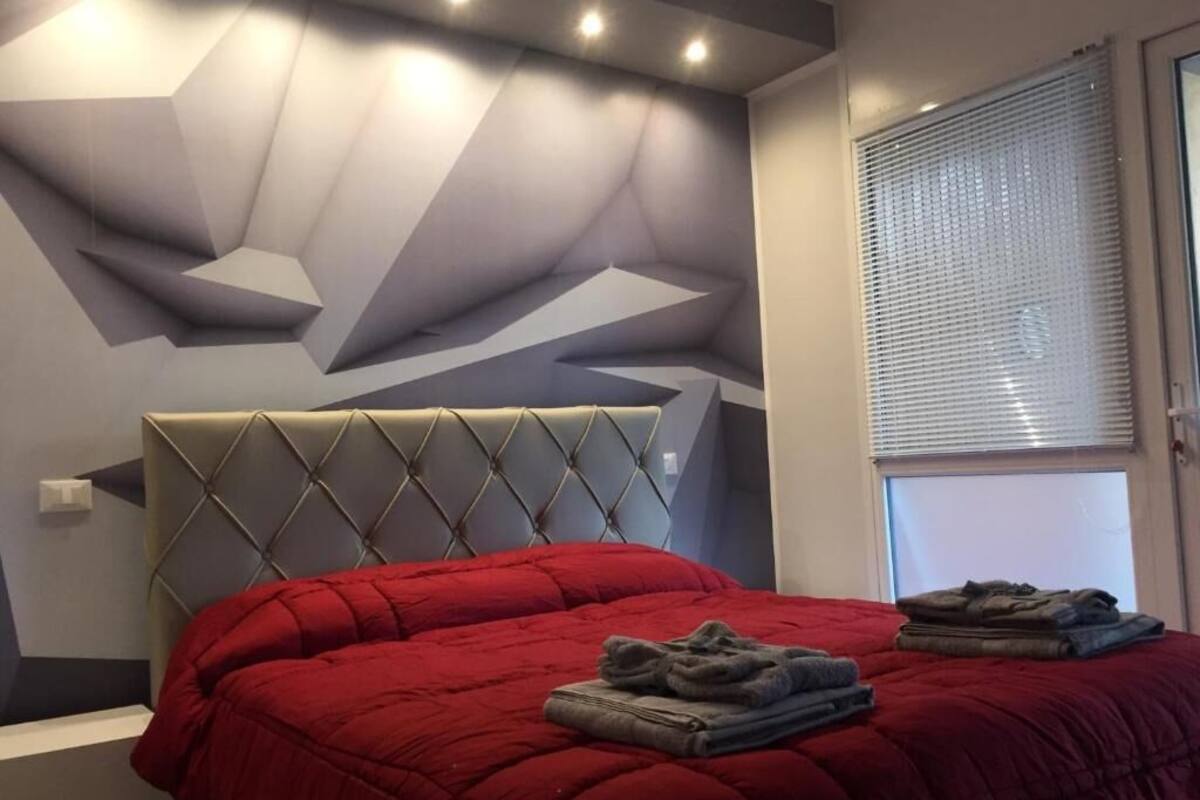 Ein Schlafzimmer mit roter Bettdecke, grauem Kopfteil und einem geometrischen Wandmuster.
