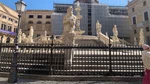 Una fuente clásica con estatuas, rodeada por una cerca de hierro forjado, frente a un edificio en construcción.