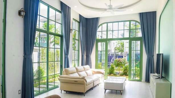 Un salon spacieux avec un canapé beige, une table basse et un téléviseur posé sur un meuble. De grandes fenêtres ornées de rideaux bleus laissent entrer la lumière naturelle et offrent une vue sur l’extérieur.