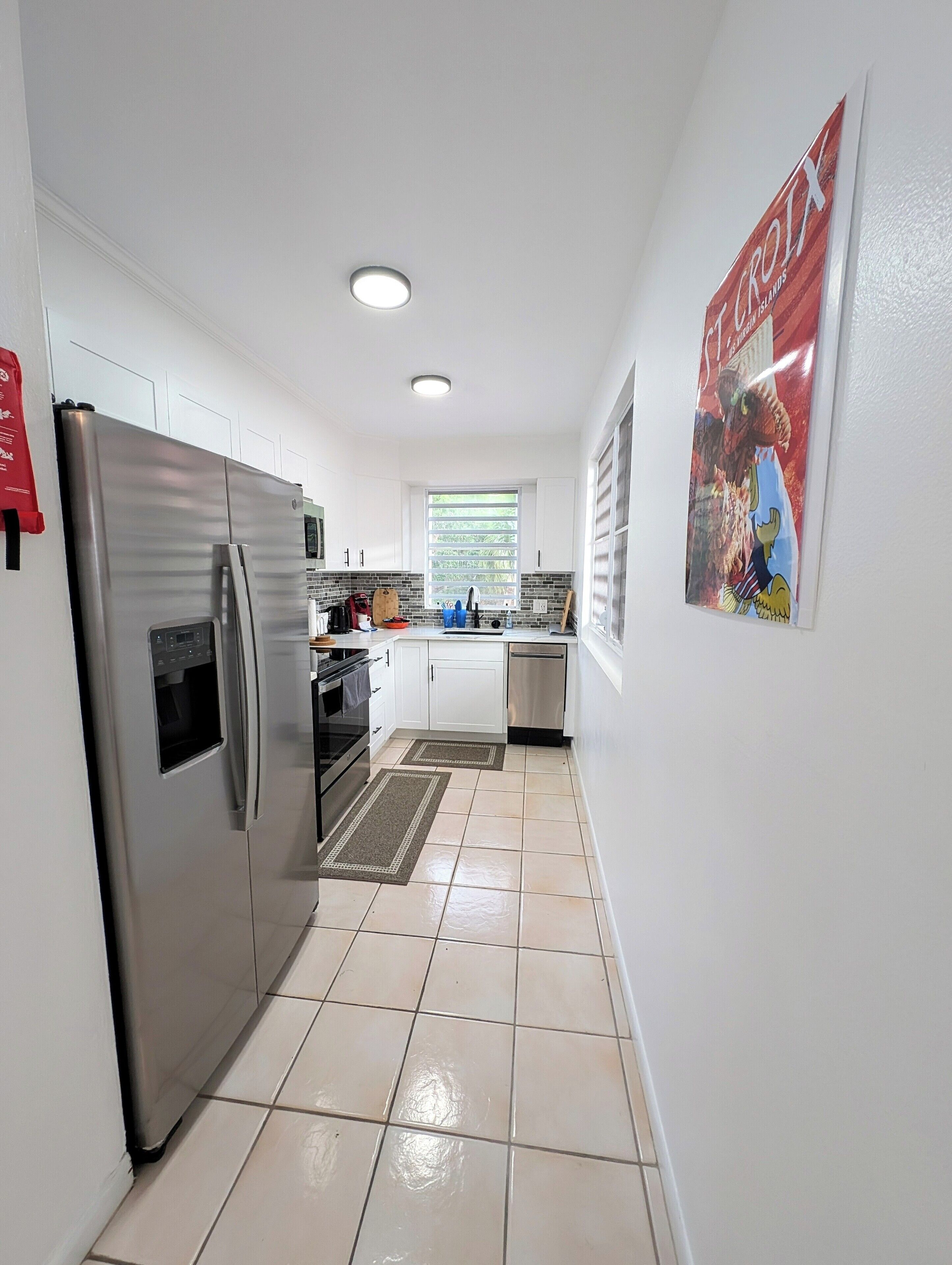 Una cocina moderna con armarios blancos, un refrigerador de acero inoxidable, horno empotrado y lavavajillas. Cuenta con una ventana con persianas, piso de baldosas y un cartel en la pared.
